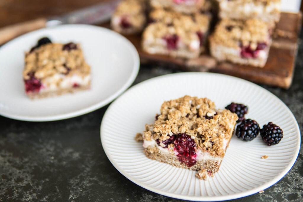 Two plates of Blackberry Cheesecake Oat Bars with bars on a cutting board.