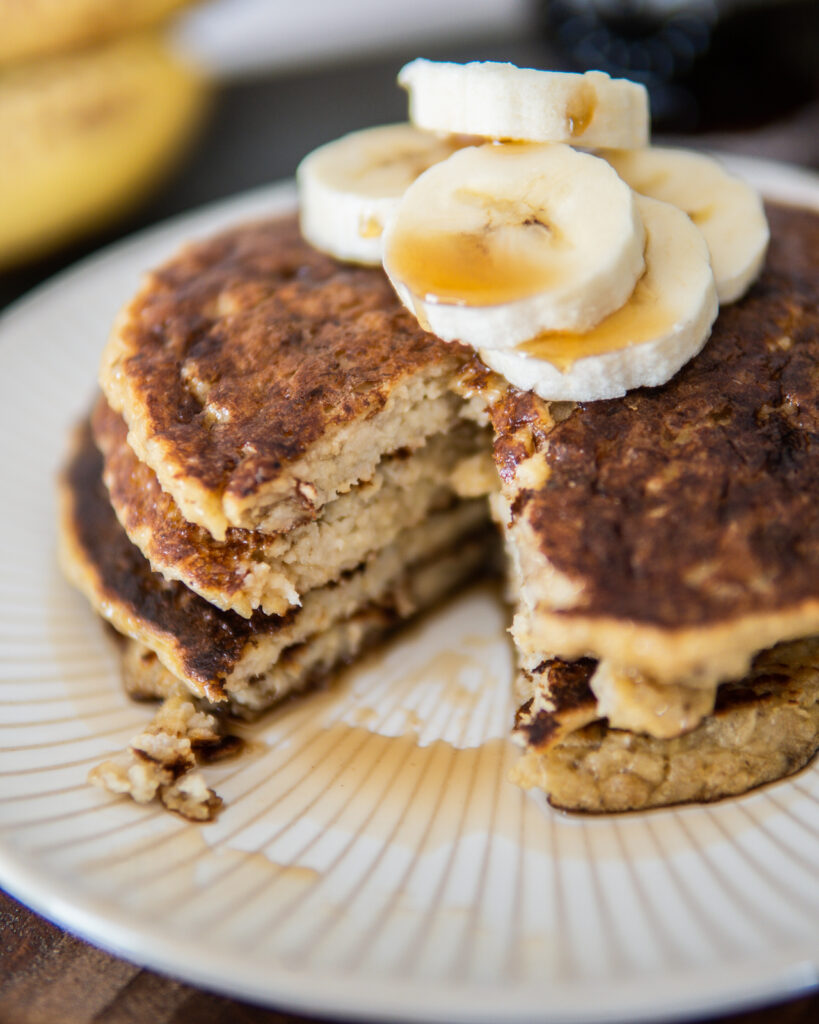 Small stack of Banana Oatmeal Pancakes with sliced bananas and maple syrup with a bite taken out.