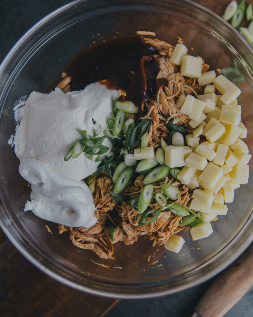 Glass bowl of ingredients: cream cheese, sour cream, pulled chicken, BBQ sauce, cheese cubes and green onions.