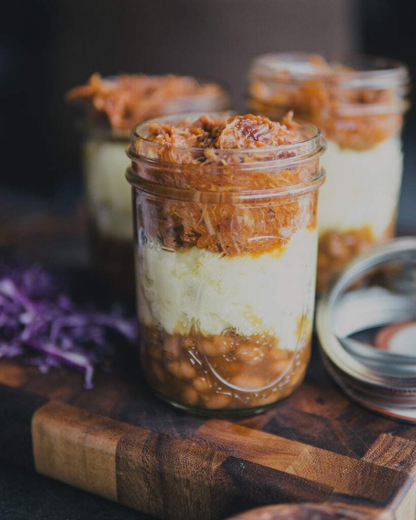 Side view of a mason jar filled with pressure cooked mashed potatoes, baked beans and Traeger smoked pulled pork. 